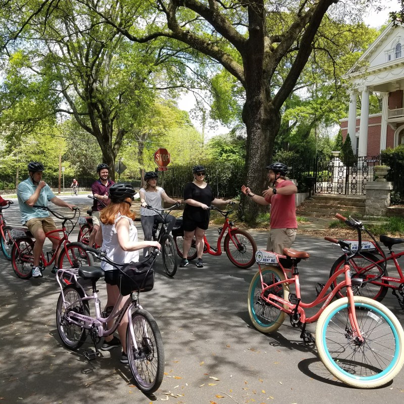 Group of people on bikes stopped on a tree-lined street, listening to a guide.