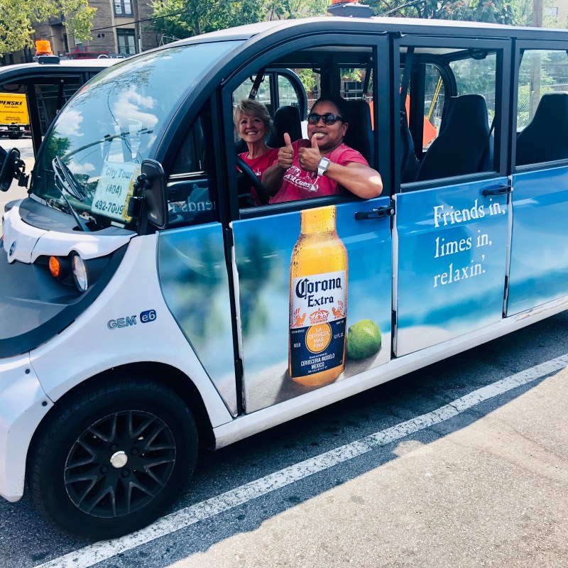 Electric car with Corona ad parked, two women inside giving thumbs up.